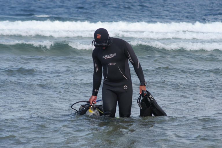 A scuba diver in a wetsuit emerges from the ocean, carrying diving equipment.