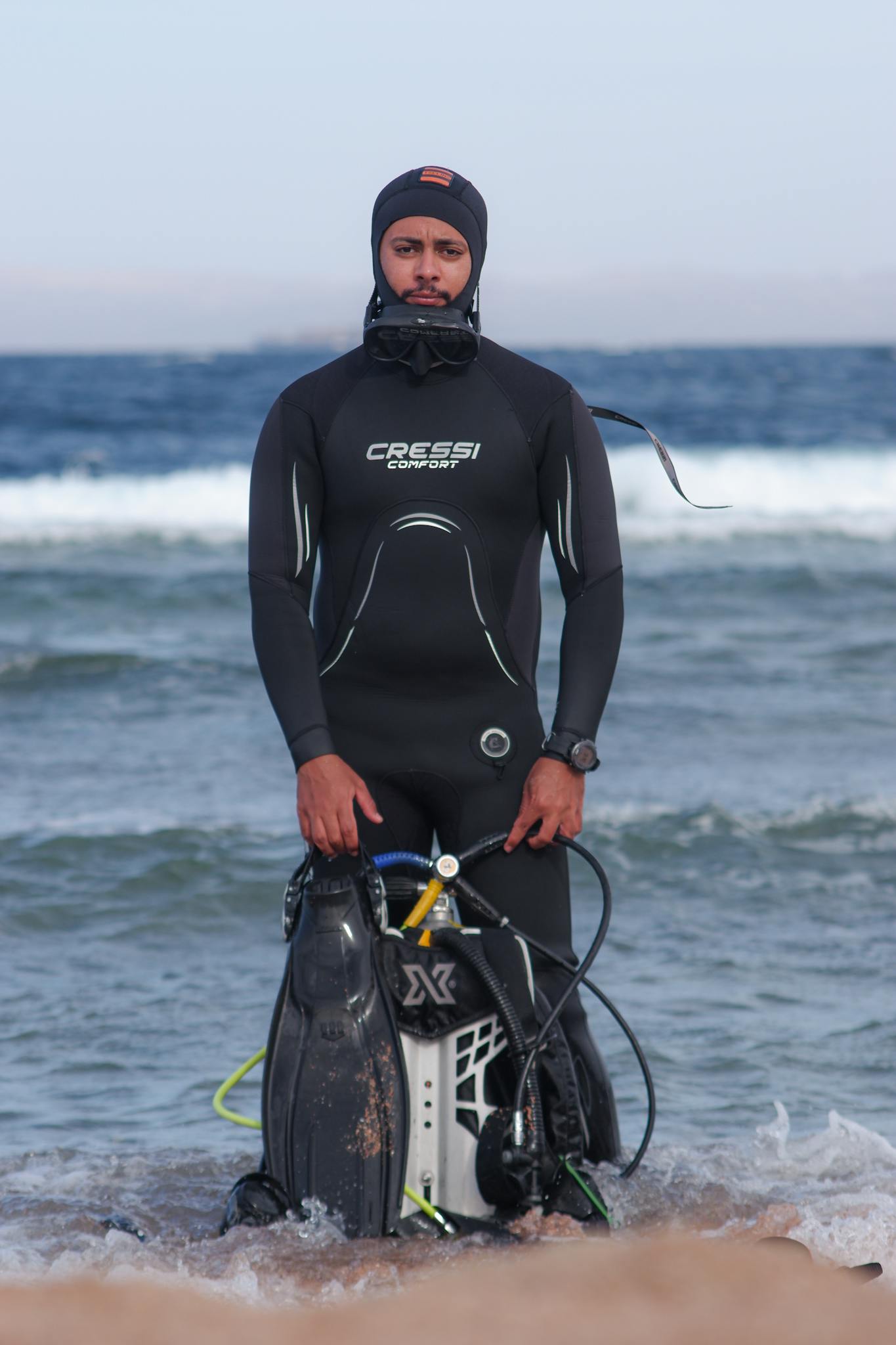 Scuba diver in wetsuit preparing for a dive in the ocean, holding diving equipment on a beach.